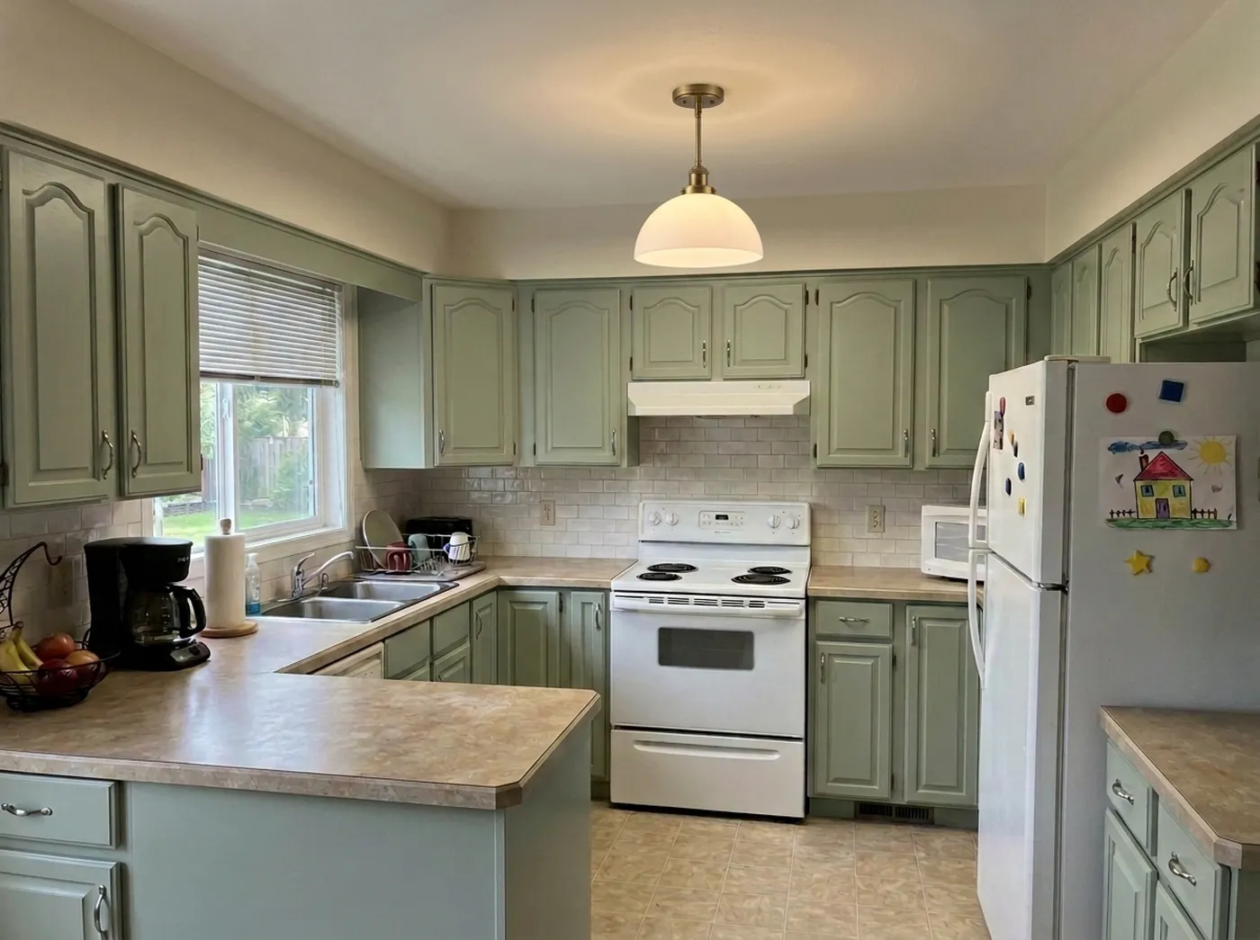 The same kitchen after: white tile backsplash, sage cabinets, and a pendant light, with everything else unchanged