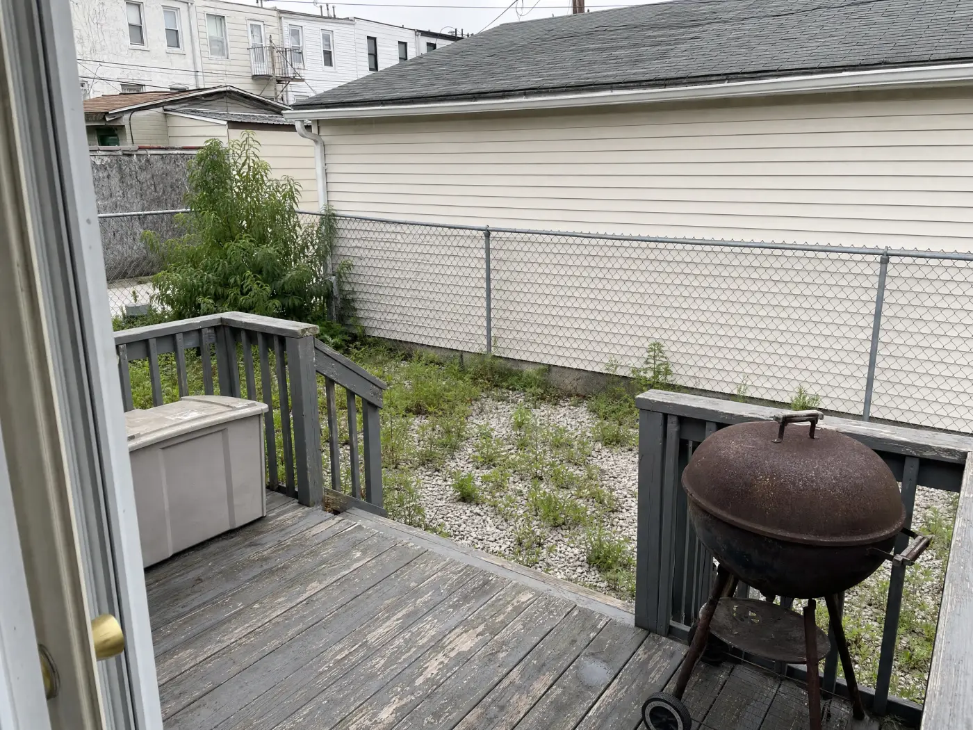 A small city backyard seen from the back door: weathered grey deck, a plastic storage box, a rusty kettle grill, weedy gravel, and a chain-link fence against the neighbor's garage