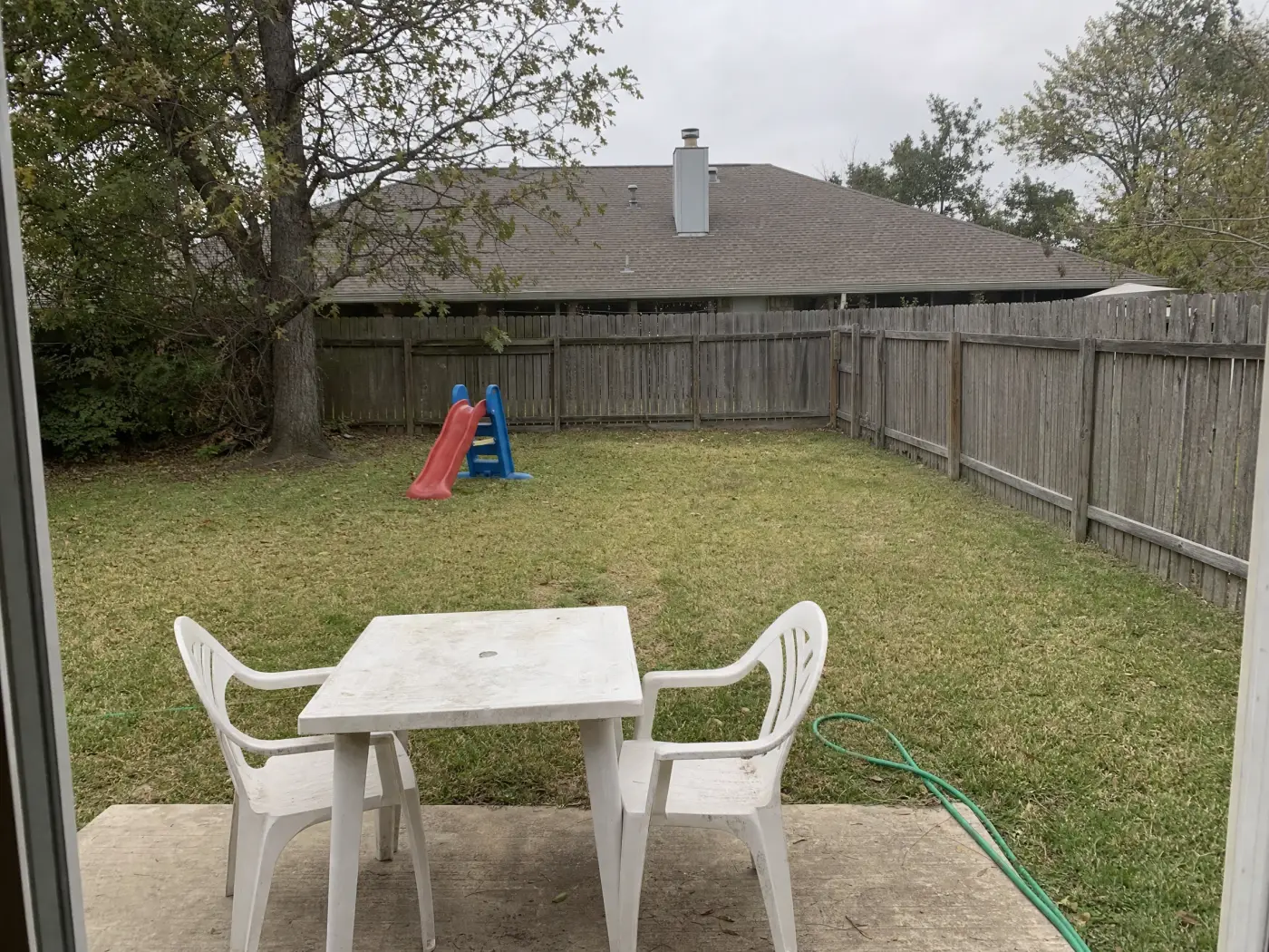 A plain family backyard seen from the back door: a bare concrete slab with a plastic table and chairs, a garden hose, a flat lawn with a child's slide, a big tree in the corner, and a weathered wooden fence, the starting point for AI Ideas in Idori