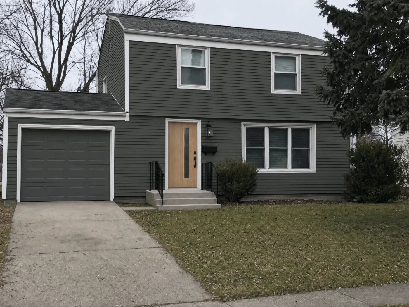 The same house after a Spot Edit repaint in Idori: deep sage green siding and garage door, the same white window trim, a white oak front door with a glass strip, a black lantern, new steps with a black railing, and the same roof, lawn, and spruce tree