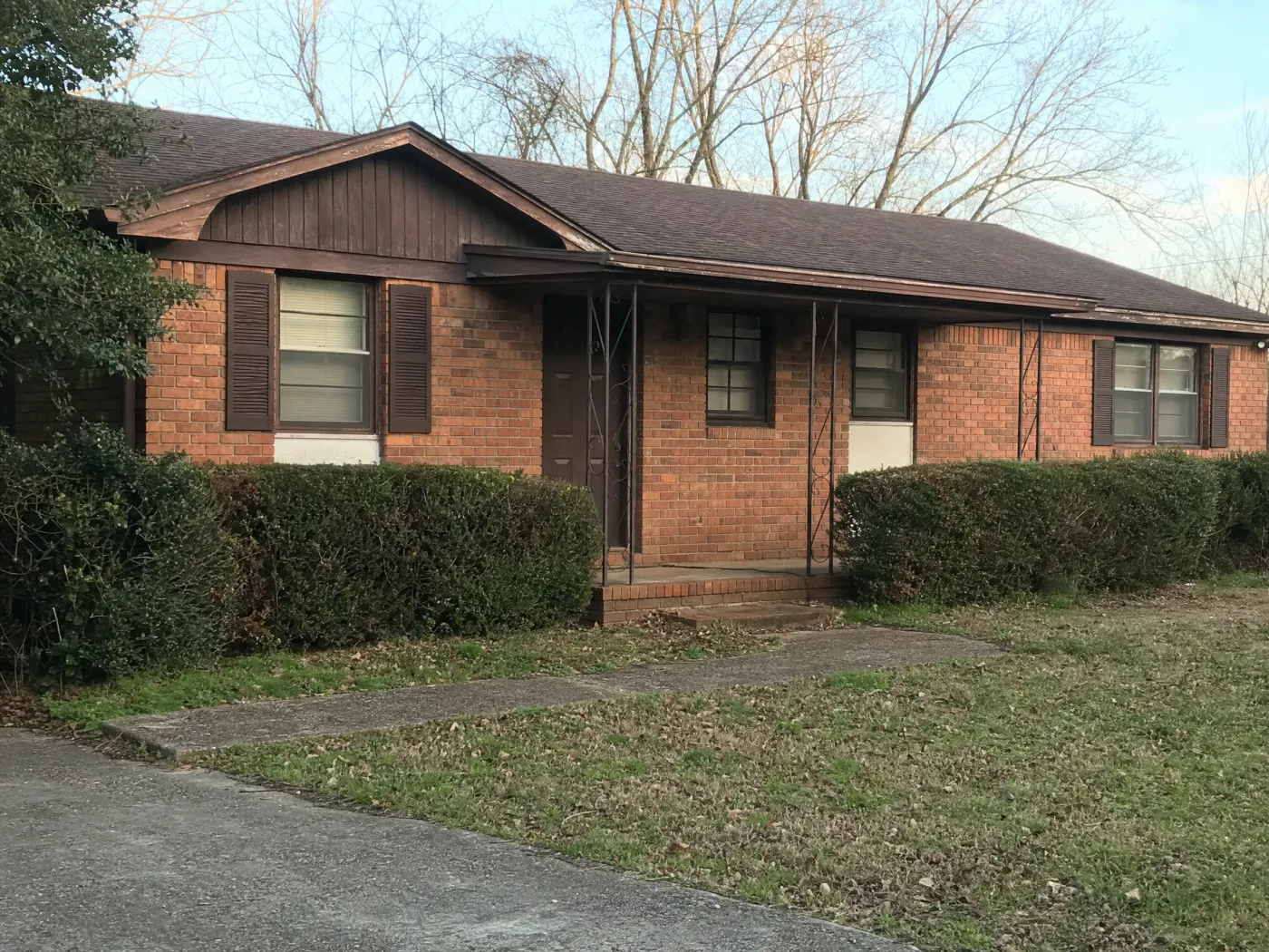 A 1970s single-story brick ranch house seen from the driveway: orange-red brick, dark brown gable boards and trim, louvered brown shutters, a thin metal porch, overgrown boxwoods, and a patchy lawn