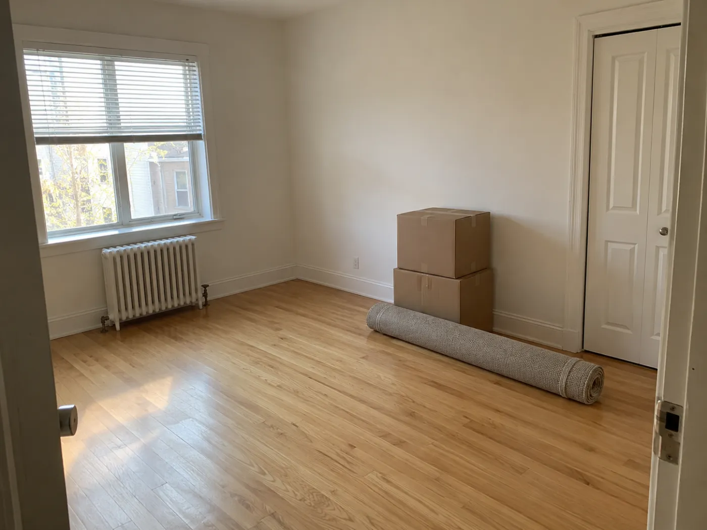 An empty bedroom on moving day: bare oak floor, a radiator under the window, moving boxes, and a rolled-up rug by the closet